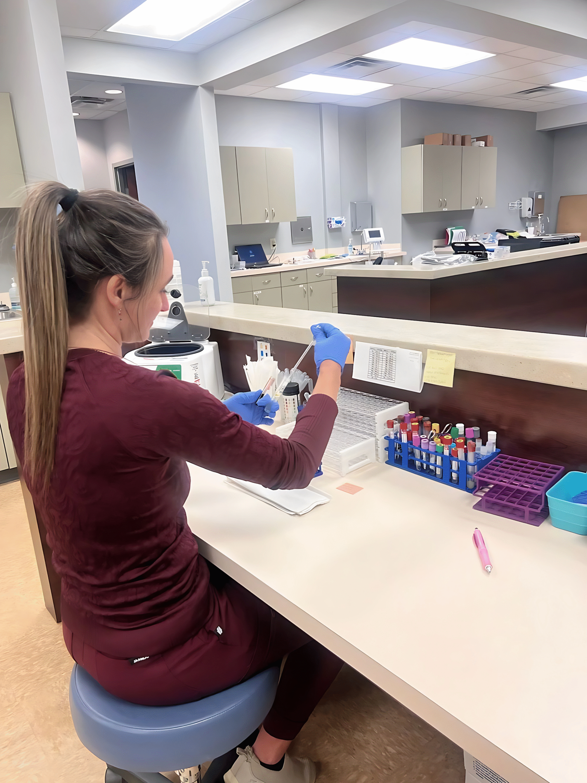 A woman in medical scrubs and gloves sits at a lab workstation, carefully handling test tubes and paperwork in a clinical setting, showing dedication to every detail about patient care.