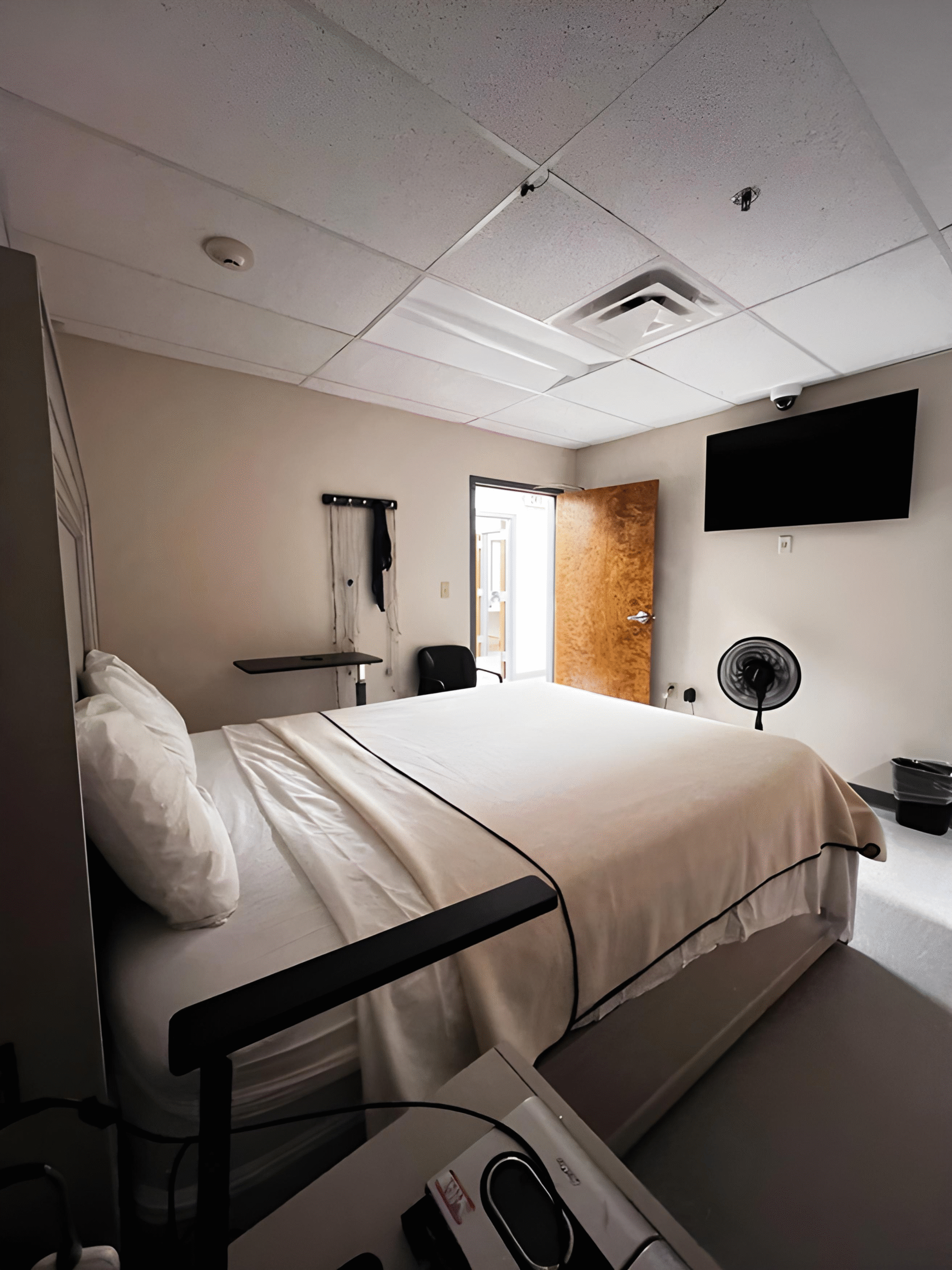 Our facility offers a small, clean room with a neatly made bed, wall-mounted TV, open door, fan, and minimal furnishings.