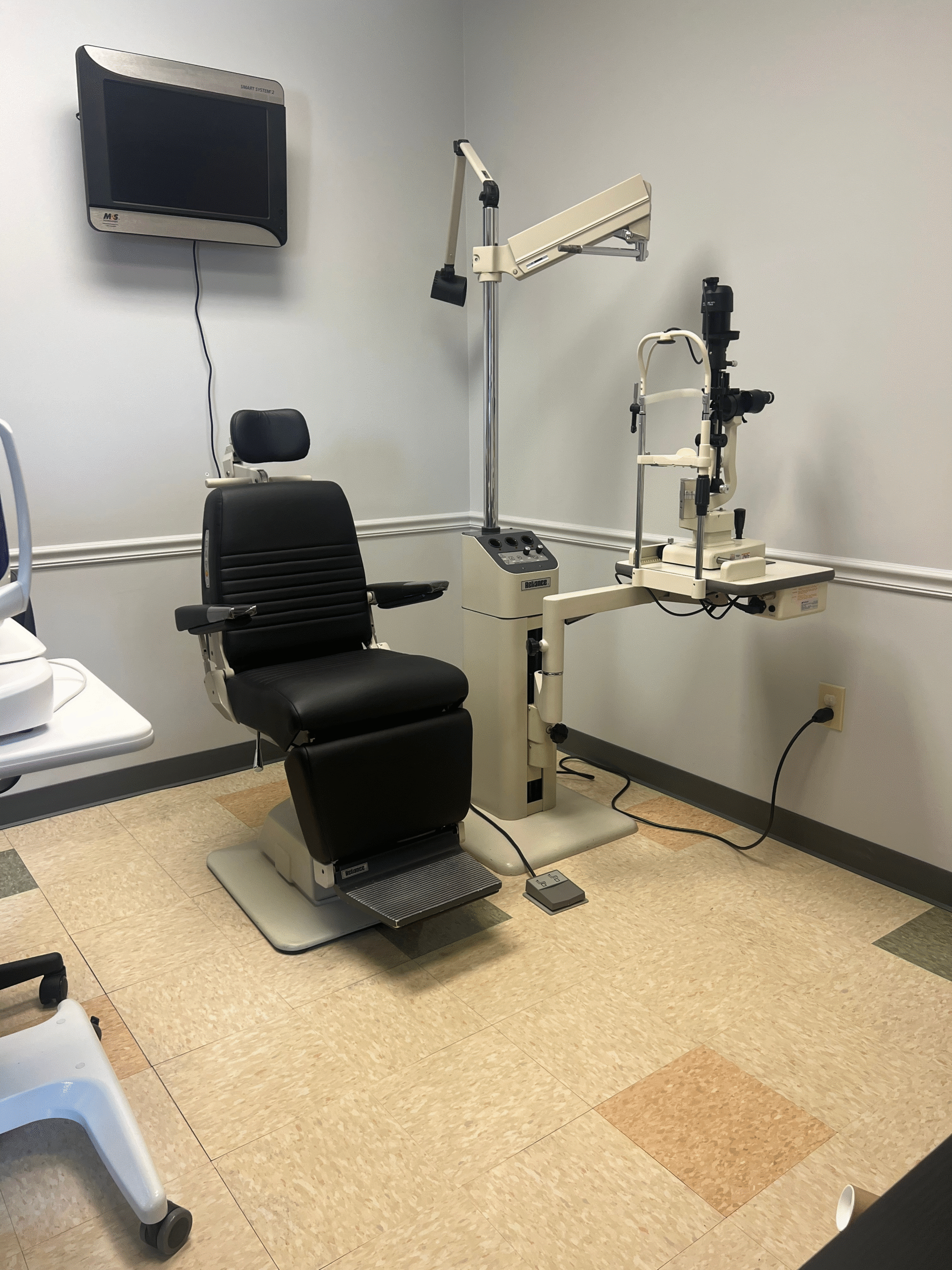 Our facility features an exam room with an ophthalmic chair, slit lamp, and a TV mounted on the wall, designed for comprehensive eye examinations.