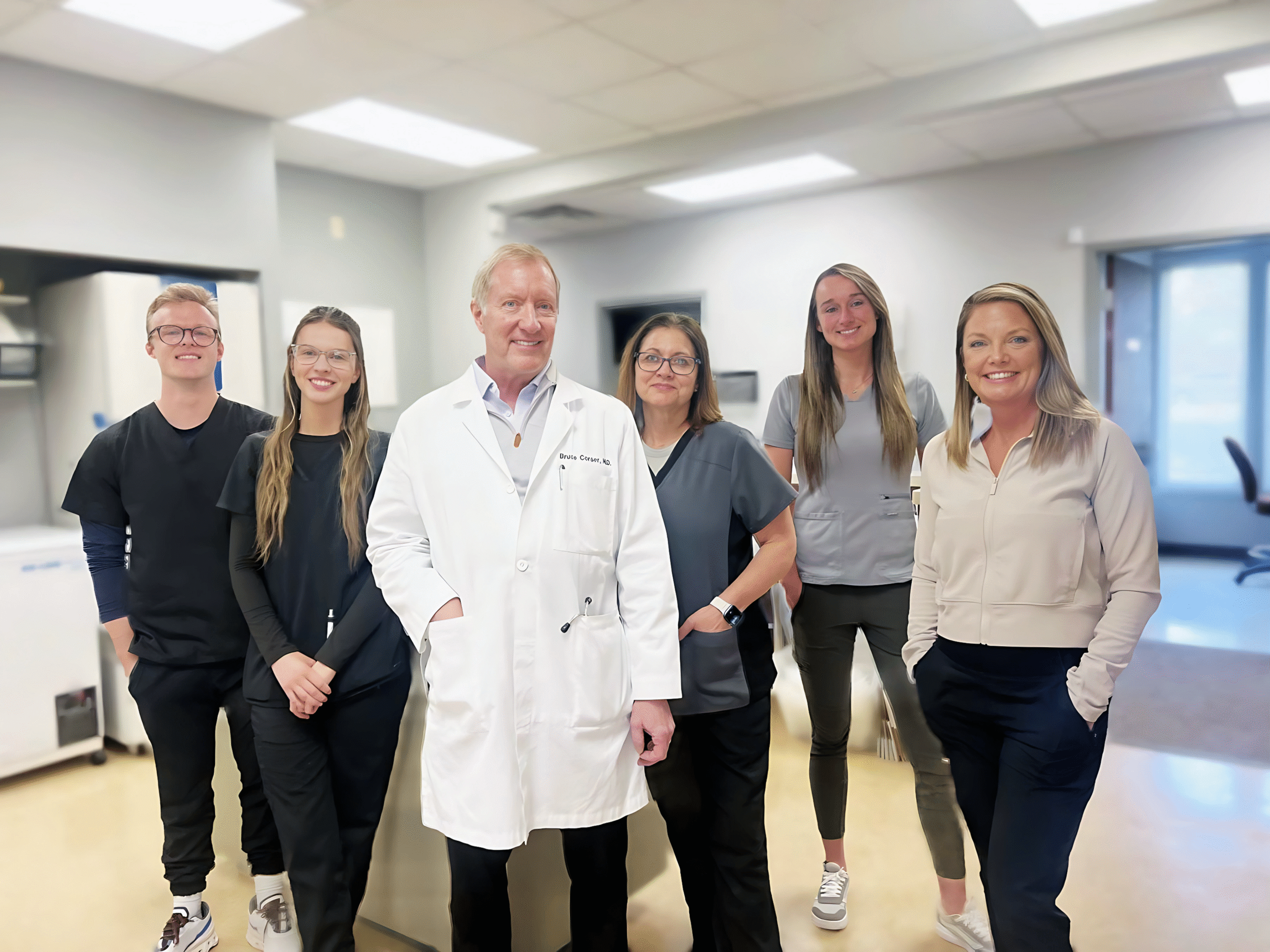 About five adults, including one person in a white lab coat and four in medical scrubs, stand indoors in a well-lit clinical setting.