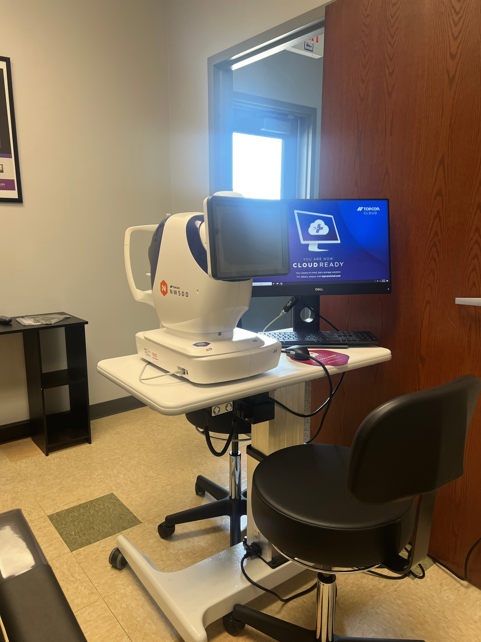 A vision screening machine sits on a rolling cart next to a computer monitor and office chair in a small exam room at our facility.