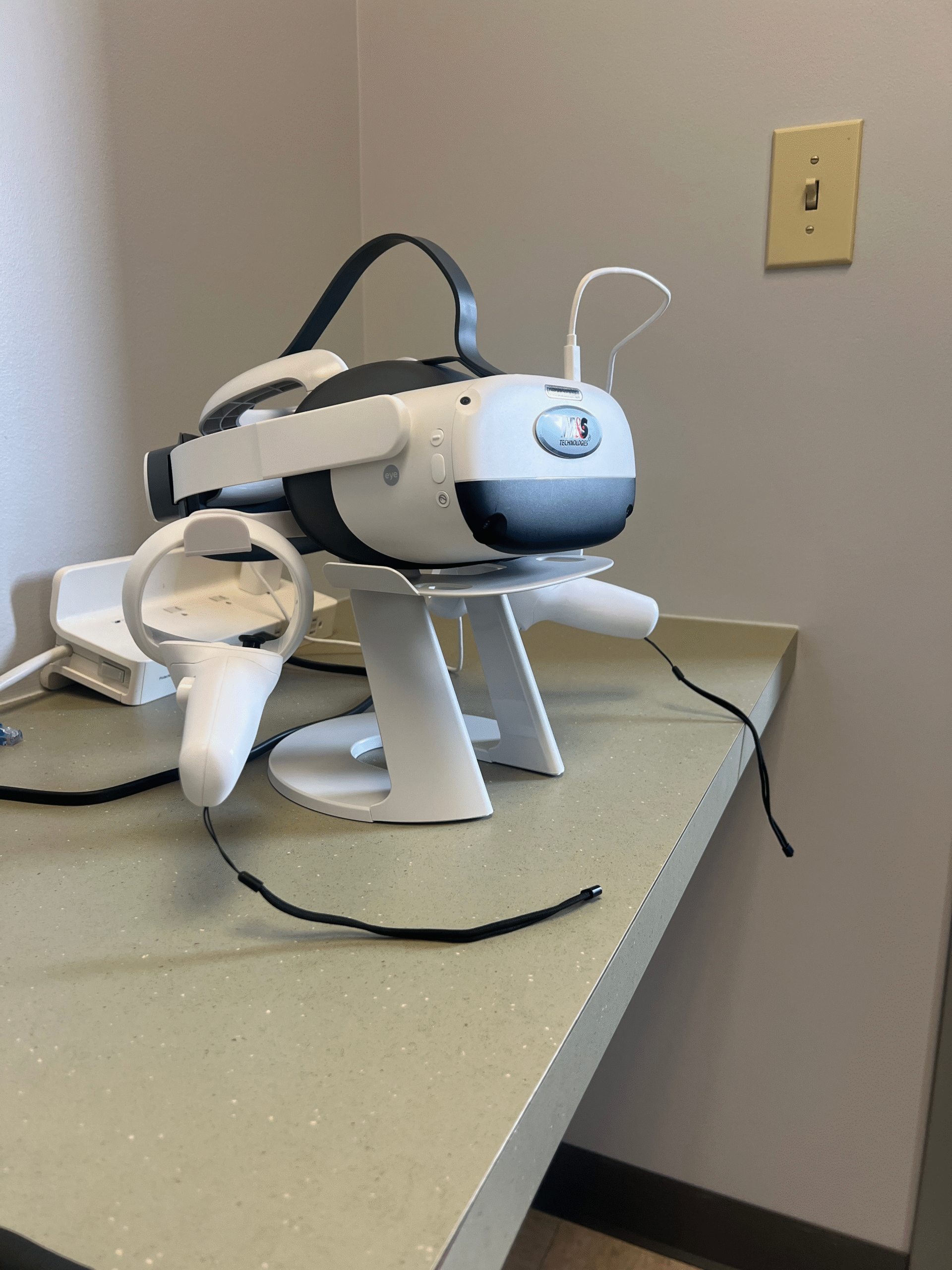 A virtual reality headset with attached controllers sits on a charging stand on a countertop in our facility, next to a wall outlet.