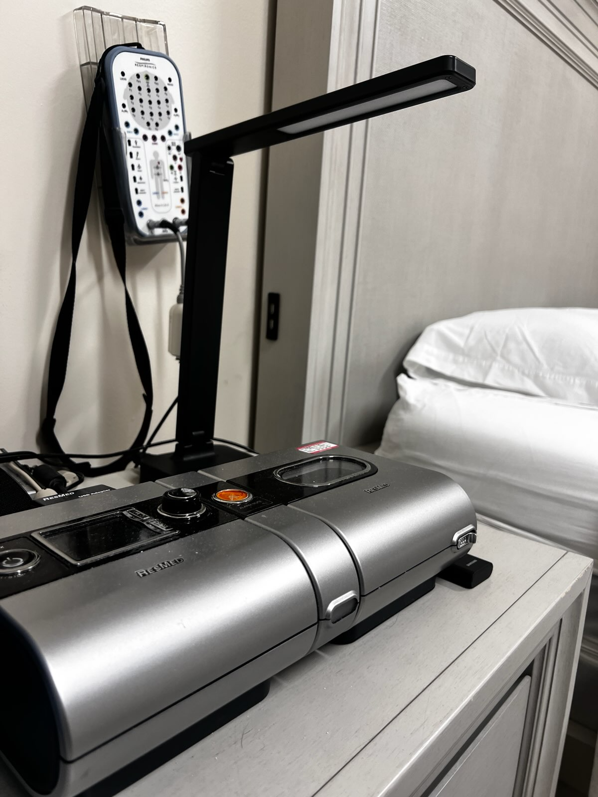 A CPAP machine and its accessories from our facility are placed on a bedside table next to a neatly made bed with white pillows.