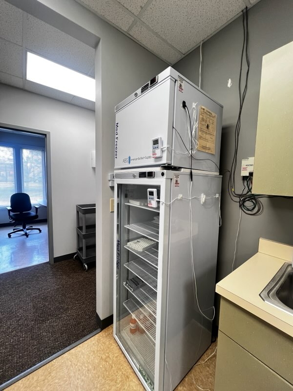 Our facility features a laboratory refrigerator and freezer with glass doors and temperature displays, standing against a gray wall near a sink and an office chair.