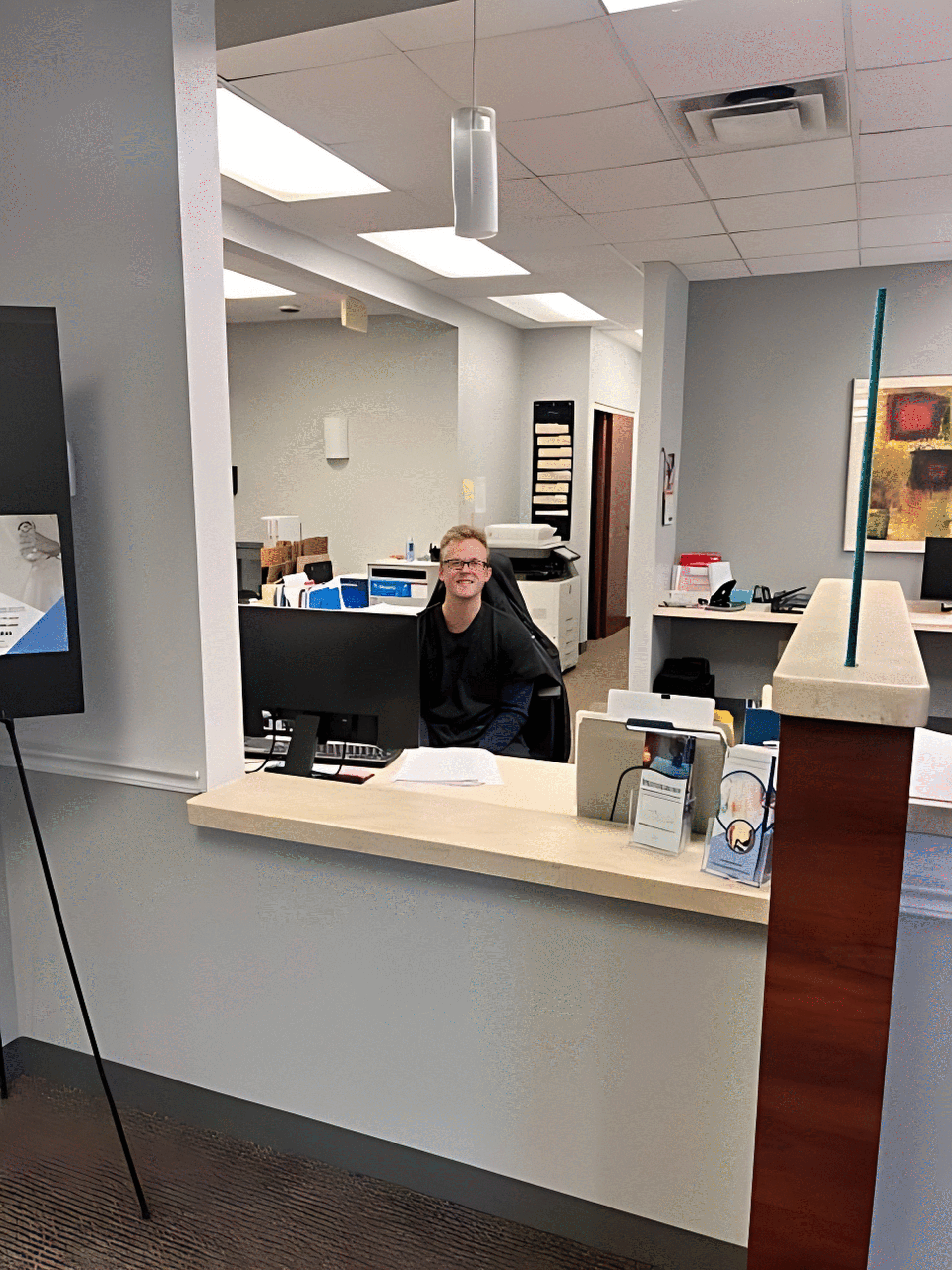 A person sits at a reception desk in an office setting, with papers, brochures, and office equipment visible on the counter and in the background, giving insight about daily administrative tasks.
