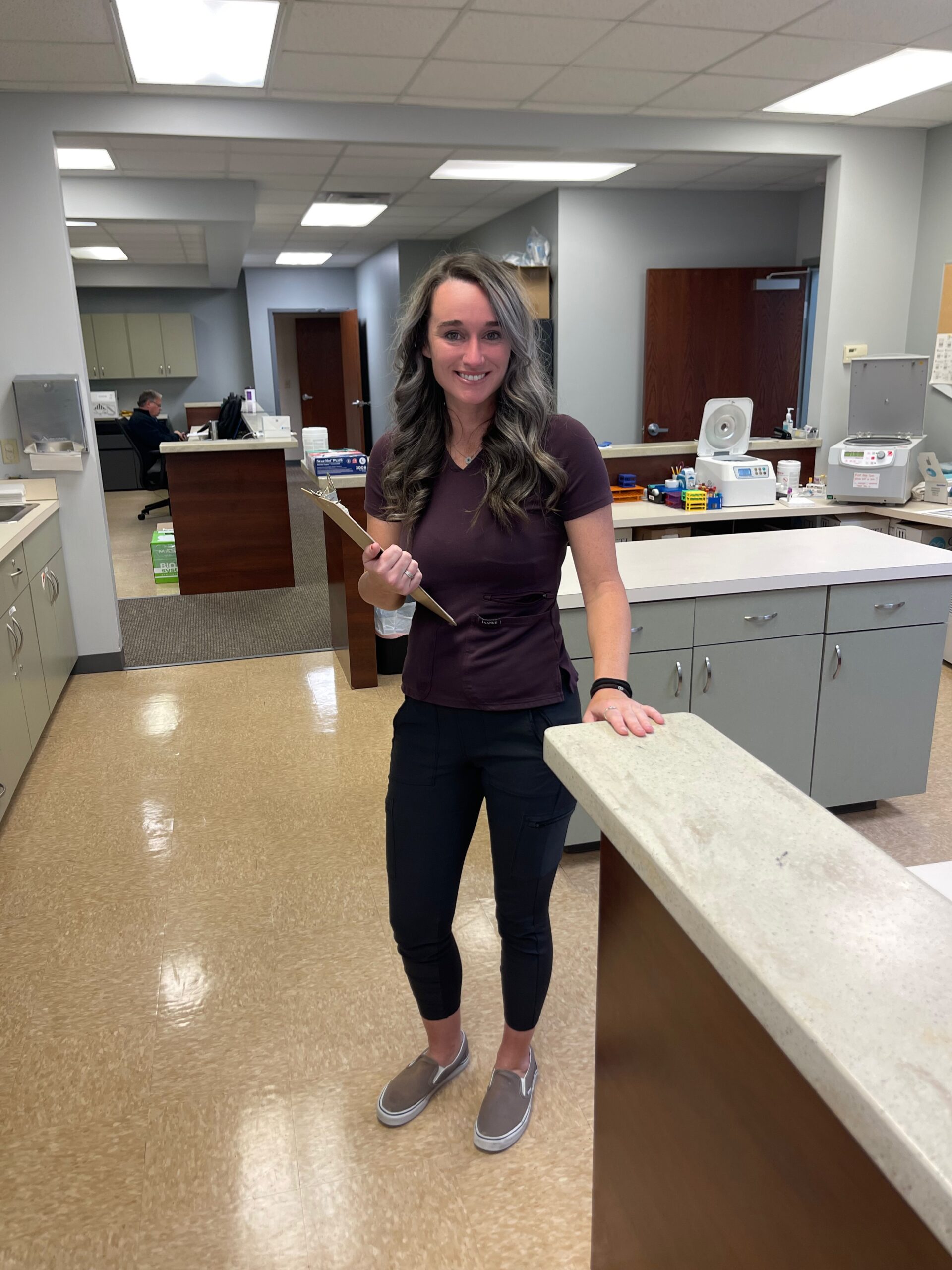 A woman in medical scrubs stands in a clinic or laboratory setting, holding a clipboard and smiling at the camera, reflecting her passion about caring for others.