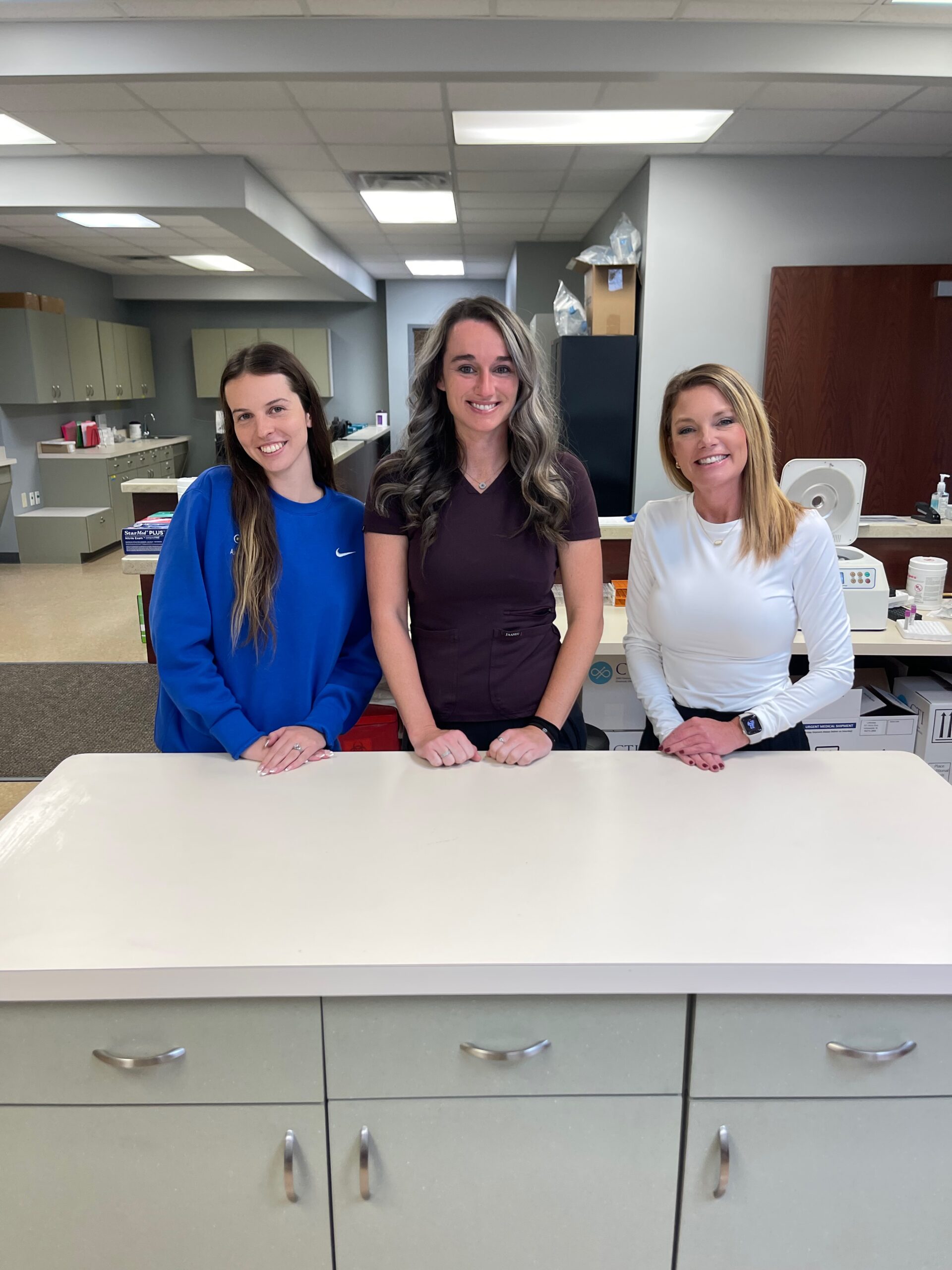 Three women stand behind a counter in an office or clinic setting, smiling at the camera, giving a warm and welcoming impression about the workplace.